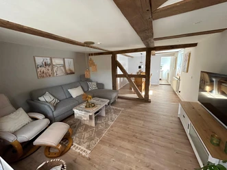 Wohnbereich der Ferienwohnung Wesertraum Lauenförde Wohnzimmer mit grauem Ecksofa, Sessel mit Fußbank, Couchtisch, gegenüber TV-Bank mit Fernseher, im hinteren Bereich Esstisch mit Stühlen, alte Holzbalken sichtbar