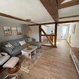 Wohnbereich der Ferienwohnung Wesertraum Lauenförde Wohnzimmer mit grauem Ecksofa, Sessel mit Fußbank, Couchtisch, gegenüber TV-Bank mit Fernseher, im hinteren Bereich Esstisch mit Stühlen, alte Holzbalken sichtbar