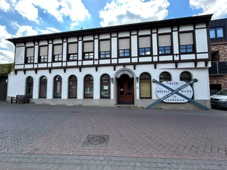 Zweistöckiges Fachwerkhaus mit Rundbogenfenstern und Eingangstür, Schild mit Schrift an der Fassade.
