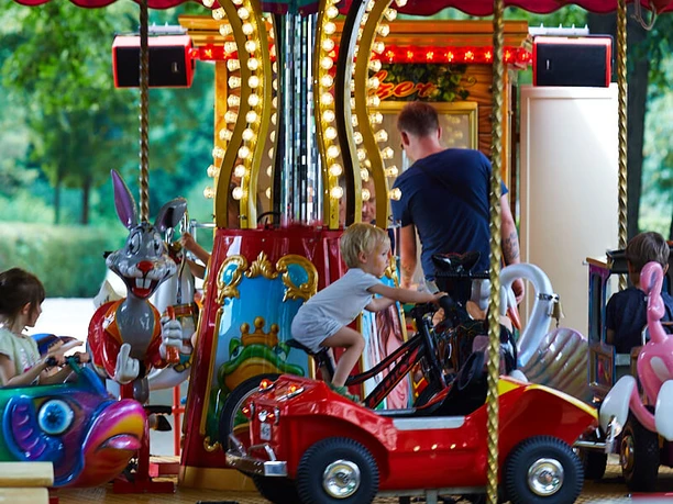 Lindenthaler-Familien-und-Veedelsfest_Krzysztof-Swider _S3-4950.jpg Ein buntes Karussell mit fröhlich lachenden Kindern, umgeben von strahlenden Lichtern und liebevoll gestalteten Figuren, dreht sich im Grünen.