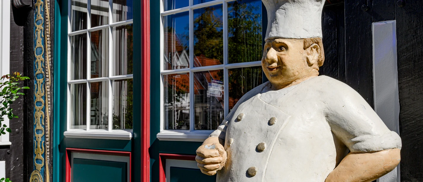 Alltagsmenschen-Koch vor Ratskeller Eine Statue eines lächelnden Kochs steht vor einem Fachwerkgebäude mit spiegelnden Fenstern.