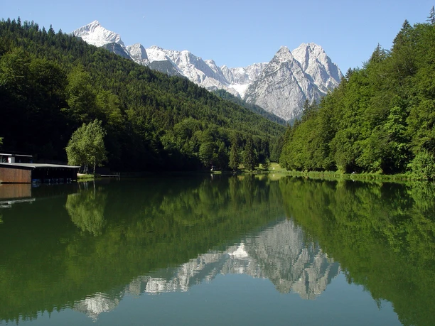 Riessersee ©GaPa Tourismus GmbHshreddhead - stock.adobe.com.jpeg Ein ruhiger Bergsee mit Waldhängen und spiegelnden Gipfeln unter klarem Himmel.