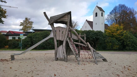Holzspielgerät mit Rutsche und Kletternetz auf Sand, dahinter eine weiße Kirche.