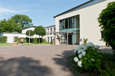 Abtei-Herstelle-Gästehaus-Betram Solcher Modernes Gästehaus mit großen Glasfenstern und Eingang, sehr gepflegte Außenanlage mit Rasenfläche, Bäumen und Hortensie.