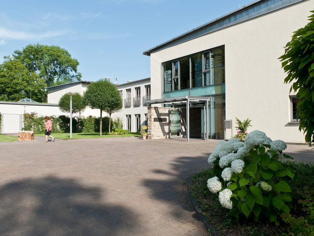 Abtei-Herstelle-Gästehaus-Betram Solcher Modernes Gästehaus mit großen Glasfenstern und Eingang, sehr gepflegte Außenanlage mit Rasenfläche, Bäumen und Hortensie.