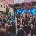 Südstadtfest Köln Eine bunte Menschenmenge feiert vor einer Bühne mit Live-Band, farbiges Konfetti in der Luft schwebend.