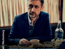 Ein Mann mit Bart sitzt vor einem Tisch in einem Restaurant, hält eine Tasse; Wasserflasche daneben.