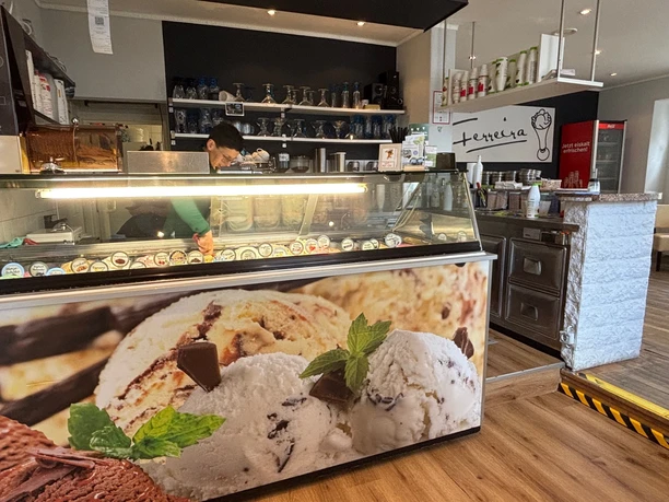 Eiscafe.jpg Eine Eisdiele mit einer Vitrine voller Eissorten und einer Person, die hinter der Theke arbeitet.