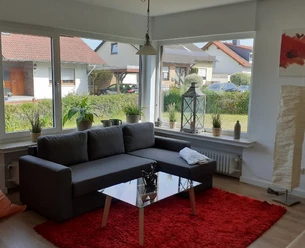 Ferienwohnung Voetter Wohnzimmer mit großer Fensterfront, Zimmerpflanzen auf der Fensterbank, grauem modernen Sofa, Glastisch