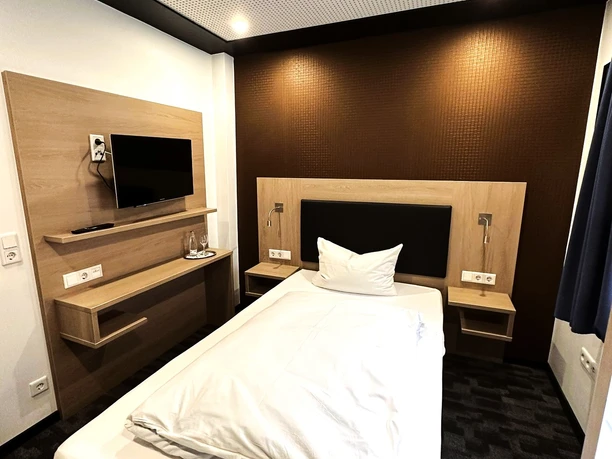 Einzelzimmer des Hotels Asado Einzelzimmer im Hotel Asado: Weißes Bett, schwarzer Boden, Leselampe, Fernseher.