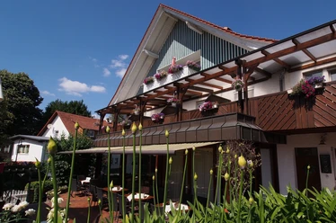 Gasthaus Kirchhoff Terrasse des Restaurants, die mit Stühlen und Tischen ausgestattet ist. Die Terrasse ist überdacht mit einer Markise. Oberhalb sind Gästezimmer mit Balkonen zu sehen, die schön bepflanzt sind.