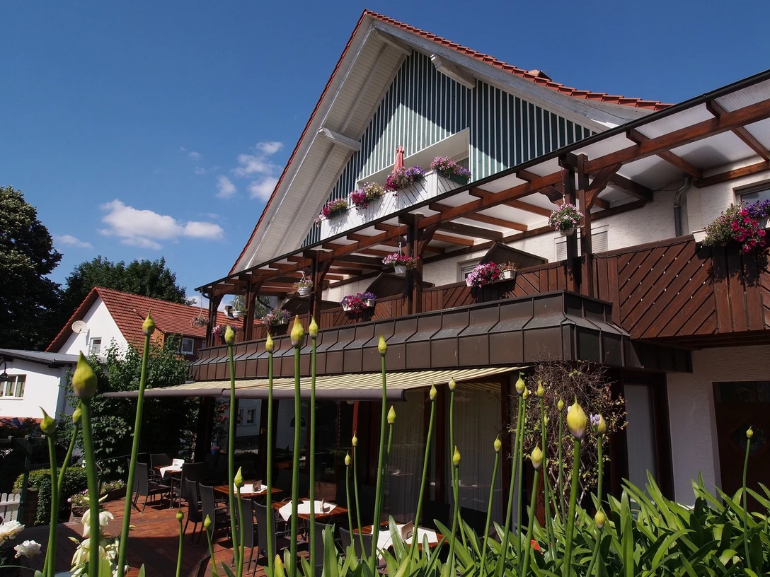 Gasthaus Kirchhoff Terrasse des Restaurants, die mit Stühlen und Tischen ausgestattet ist. Die Terrasse ist überdacht mit einer Markise. Oberhalb sind Gästezimmer mit Balkonen zu sehen, die schön bepflanzt sind.