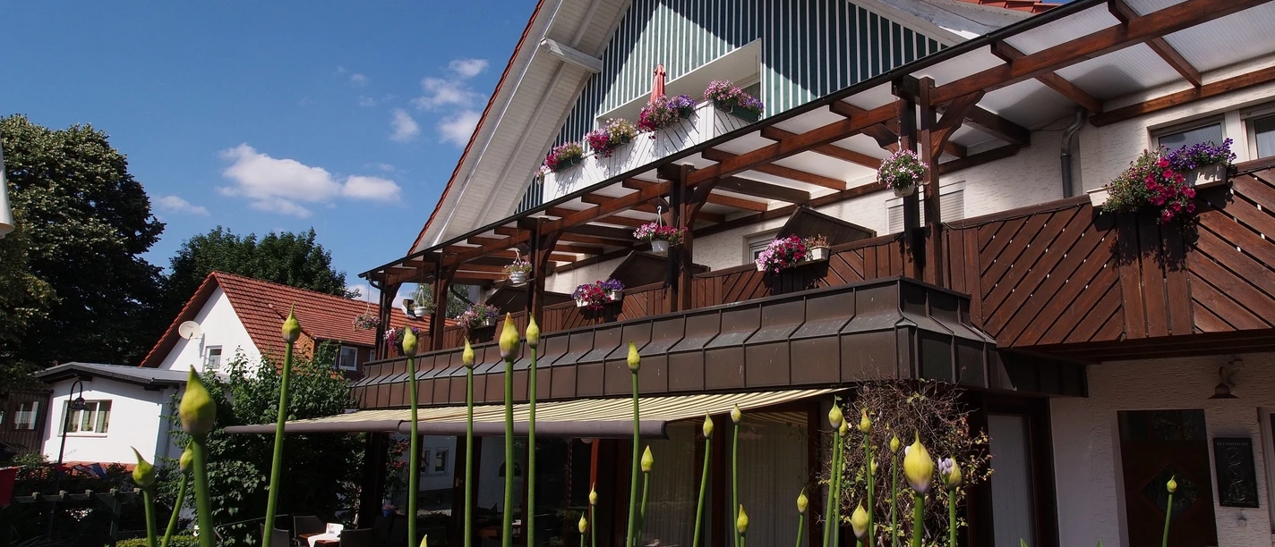 Gasthaus Kirchhoff Terrasse des Restaurants, die mit Stühlen und Tischen ausgestattet ist. Die Terrasse ist überdacht mit einer Markise. Oberhalb sind Gästezimmer mit Balkonen zu sehen, die schön bepflanzt sind.