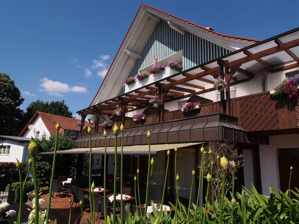 Gasthaus Kirchhoff Terrasse des Restaurants, die mit Stühlen und Tischen ausgestattet ist. Die Terrasse ist überdacht mit einer Markise. Oberhalb sind Gästezimmer mit Balkonen zu sehen, die schön bepflanzt sind.