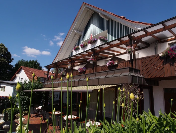 Gasthaus Kirchhoff Terrasse des Restaurants, die mit Stühlen und Tischen ausgestattet ist. Die Terrasse ist überdacht mit einer Markise. Oberhalb sind Gästezimmer mit Balkonen zu sehen, die schön bepflanzt sind.