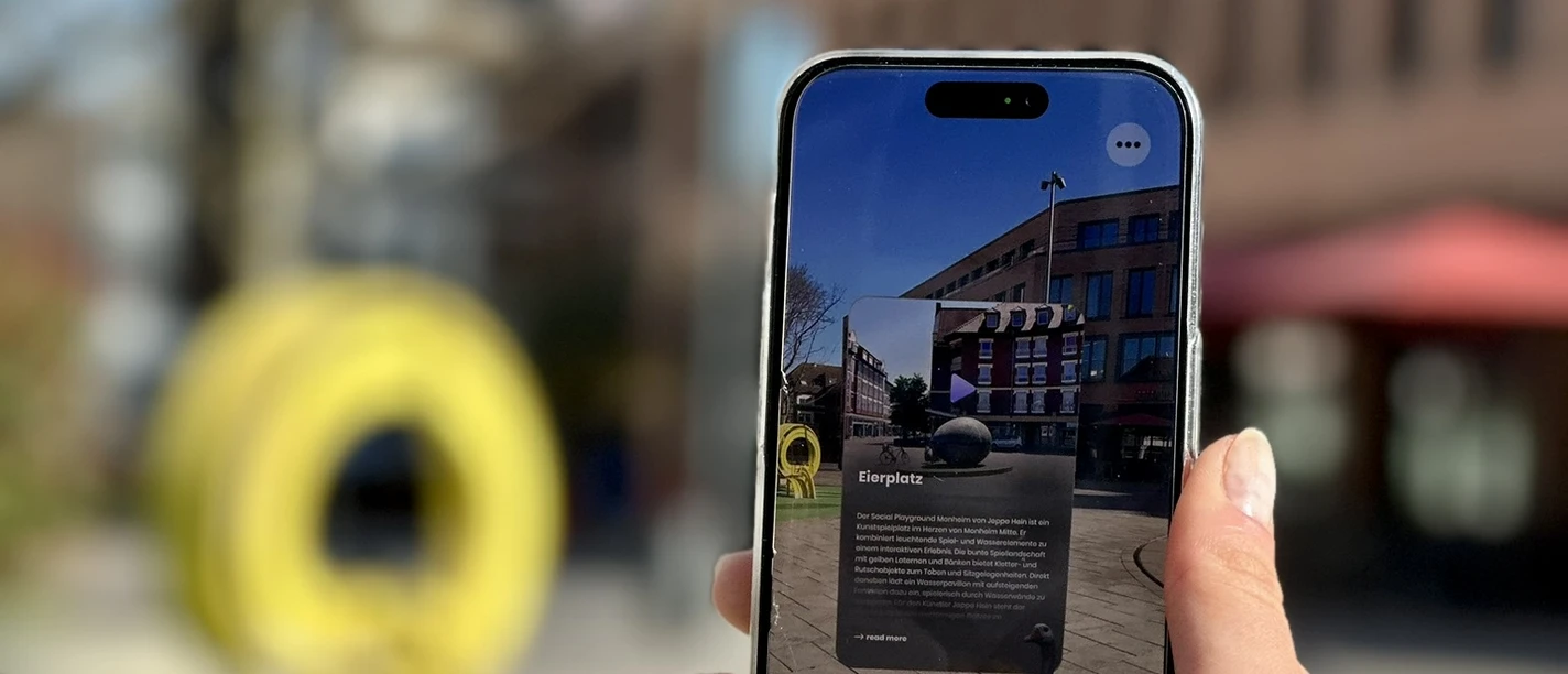 Stadtrallye.jpg Ein Smartphone wird draußen gehalten, das eine App mit Augmented Reality enthält, während im Hintergrund ein gelbes Kunstobjekt und Gebäudefronten sichtbar sind.