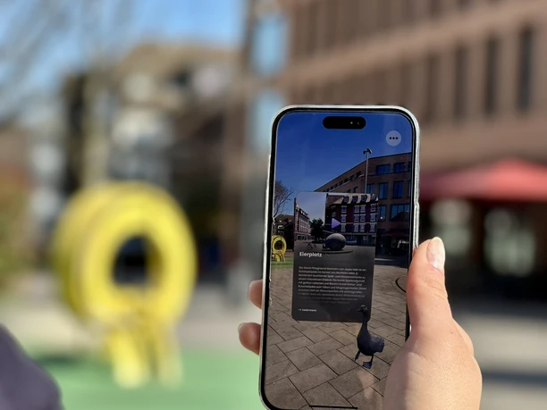 Stadtrallye.jpg Ein Smartphone wird draußen gehalten, das eine App mit Augmented Reality enthält, während im Hintergrund ein gelbes Kunstobjekt und Gebäudefronten sichtbar sind.