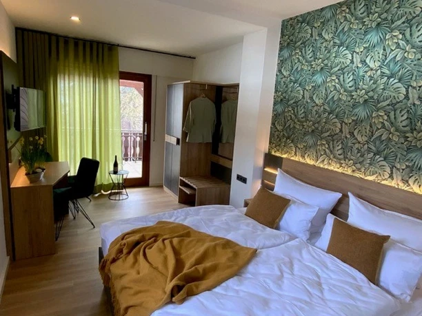 Zimmer des Gasthauses Kirchhoff Doppelzimmer mit modernem Bett aus Holz, floraler Tapete am Kopfende, Schreibtisch und Stuhl, 2-türiger Kleiderschrank, grüne Gardinen, Balkonzugang