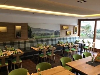 Restaurant Heimatliebe - Gasthaus Kirchhoff Restaurant Heimatliebe, zur Wand Vierertische mit jeweils zwei Stühlen mit grüner Polsterung und Sitzbank im Karodesign, an der Wand ein Landschaftsbild und Dekoration aus Holzkästen