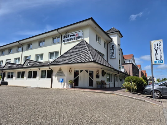 Schlossparkhotel Hof von Oldenburg Das Bild zeigt ein zweigeschossiges Hotel im klassischen Stil mit weißer Fassade und markantem dunkelgrauem Dach. Ein gepflegter Parkplatz mit einem deutlich sichtbaren Behindertenparkplatz ist ebenfalls vorhanden. Der klare Himmel und die gut gepflegte Außenanlage vermitteln einen einladenden Eindruck.
