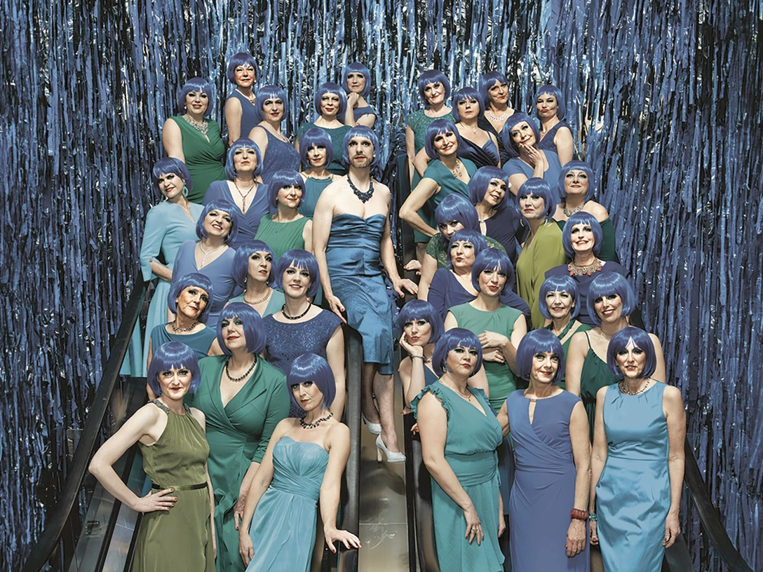 damenlikorchor-c-thomas-rusch-quer-cmyk_rgb.jpg Gruppe in grünen und blauen Kleidern mit blauen Perücken auf einer Rolltreppe vor Glitzerkulisse