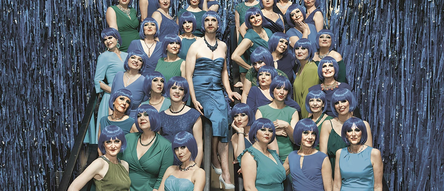 damenlikorchor-c-thomas-rusch-quer-cmyk_rgb.jpg Gruppe in grünen und blauen Kleidern mit blauen Perücken auf einer Rolltreppe vor Glitzerkulisse
