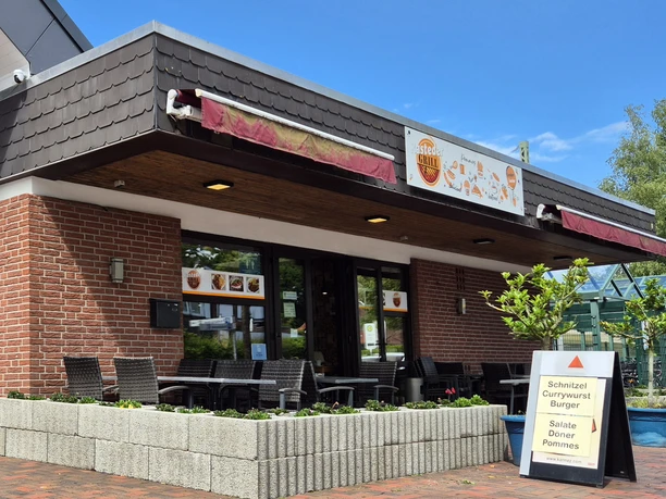 Aussenansicht Rasteder Grill Ein kleines, rotes Backsteingebäude mit Terrasse und überdachten Sitzplätzen wird von einem Schild mit Speiseangebot flankiert.