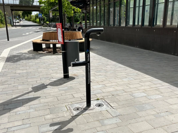 Trinkwasserbrunnen auf einem Bahnhofsplatz mit Sitzbank im Hintergrund, geeignet für durstige Reisende.