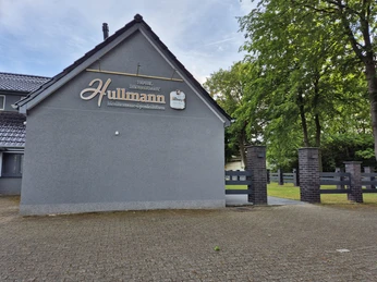 Restaunrant Hullmann Seitenansicht Graues Gebäude mit giebelseitigem Firmenschild "Hullmann Fleischerei-Spezialitäten", umgeben von Bäumen und gepflastertem Vorplatz.