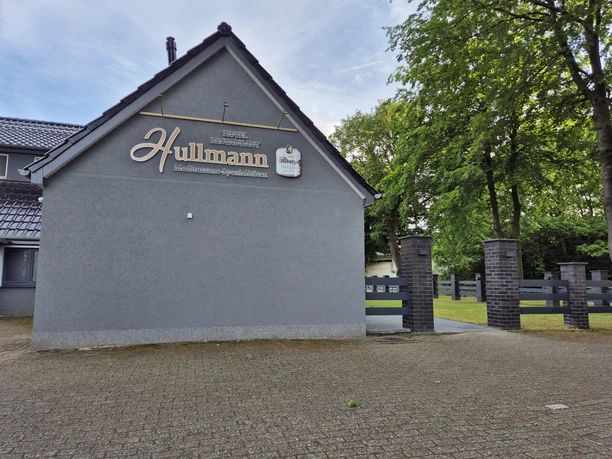 Restaunrant Hullmann Seitenansicht Graues Gebäude mit giebelseitigem Firmenschild "Hullmann Fleischerei-Spezialitäten", umgeben von Bäumen und gepflastertem Vorplatz.