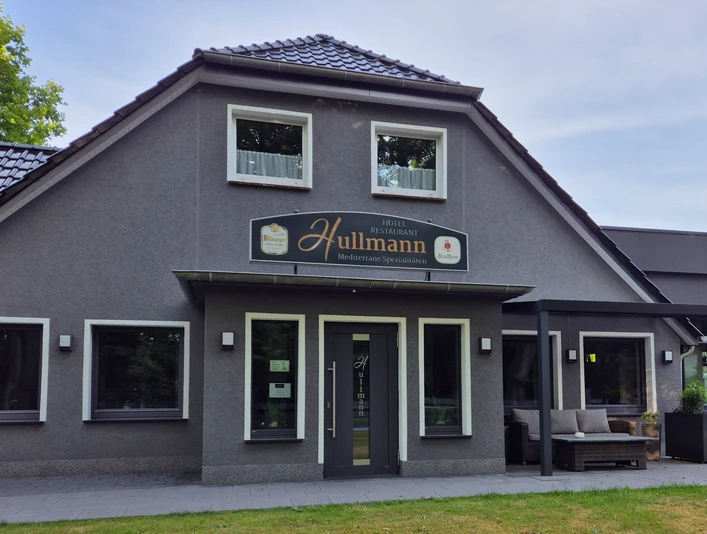 Restaurant Hullmann Aussenansicht Das Bild zeigt ein modernes Gebäude mit grauer Fassade und zahlreichen Fenstern. Vor dem Gebäude hängt ein Schild mit der Aufschrift "Hotel & Restaurant Hullmann". Rechts neben dem Eingang befindet sich eine überdachte Sitzlounge. Der Rasen im Vordergrund ist gepflegt und bietet eine grüne Begrenzung zum Gehweg.