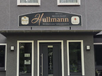 Restaurant Hullmann Eingang Das Bild zeigt die Fassade des Hotels Hullmann mit ansprechendem, modernem Design in einem schlichten Grauton, unterbrochen durch große Fenster und einer Eingangstür, auf der "Hullmann" steht. Beschriftungen an der Fassade verweisen auf das Restaurant mit mediterranen Spezialitäten. Der gepflegte Eingangsbereich wird von einer sauberen Ziersteinmauer eingefasst, was dem Gebäude ein einladendes Erscheinungsbild verleiht.