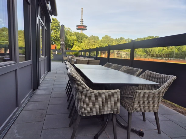 Restaurant Hullmann außen Terasse Auf dem Bild ist eine moderne Terrasse mit einem großen Tisch und mehreren geflochtenen Stühlen zu sehen, die zum Verweilen in einem einladenden Ambiente einladen. Im Hintergrund ist ein Fernsehturm vor einer grünen Baumkulisse sichtbar, was der Szene eine urbane Note verleiht.