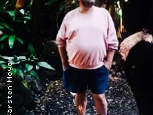 Ein Mann in lässiger Kleidung steht in einem dichten, bewaldeten Umfeld, umgeben von Laub.