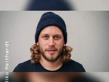 Ein Mann mit mittellangem Haar und blauem Wollmütze blickt freundlich in die Kamera vor neutralem Hintergrund.