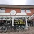 Aussenansicht Haase's Bikefactory Ein Fahrradgeschäft mit dem Namen "Haase's Bikefactory" zeigt eine Reihe von Fahrrädern vor seinem Eingang. Ein Schild weist auf einen Rabatt von bis zu 40% auf E-Bikes hin. Die Fassade besteht aus rotem Backstein und großen Fenstern. Ein blühender Baum rechts ergänzt das freundliche Ambiente.