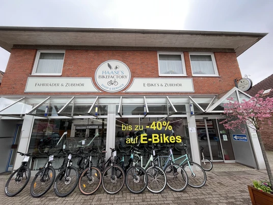Aussenansicht Haase's Bikefactory Ein Fahrradgeschäft mit dem Namen "Haase's Bikefactory" zeigt eine Reihe von Fahrrädern vor seinem Eingang. Ein Schild weist auf einen Rabatt von bis zu 40% auf E-Bikes hin. Die Fassade besteht aus rotem Backstein und großen Fenstern. Ein blühender Baum rechts ergänzt das freundliche Ambiente.