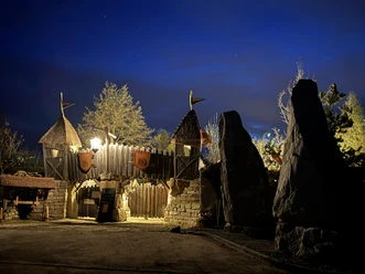 Bastei Erlebnis Steinreich Holztor mit Türmen und Fahnen, beleuchtet bei Nacht, umgeben von Bäumen und großen Steinen, vor einem tiefblauen Himmel.
