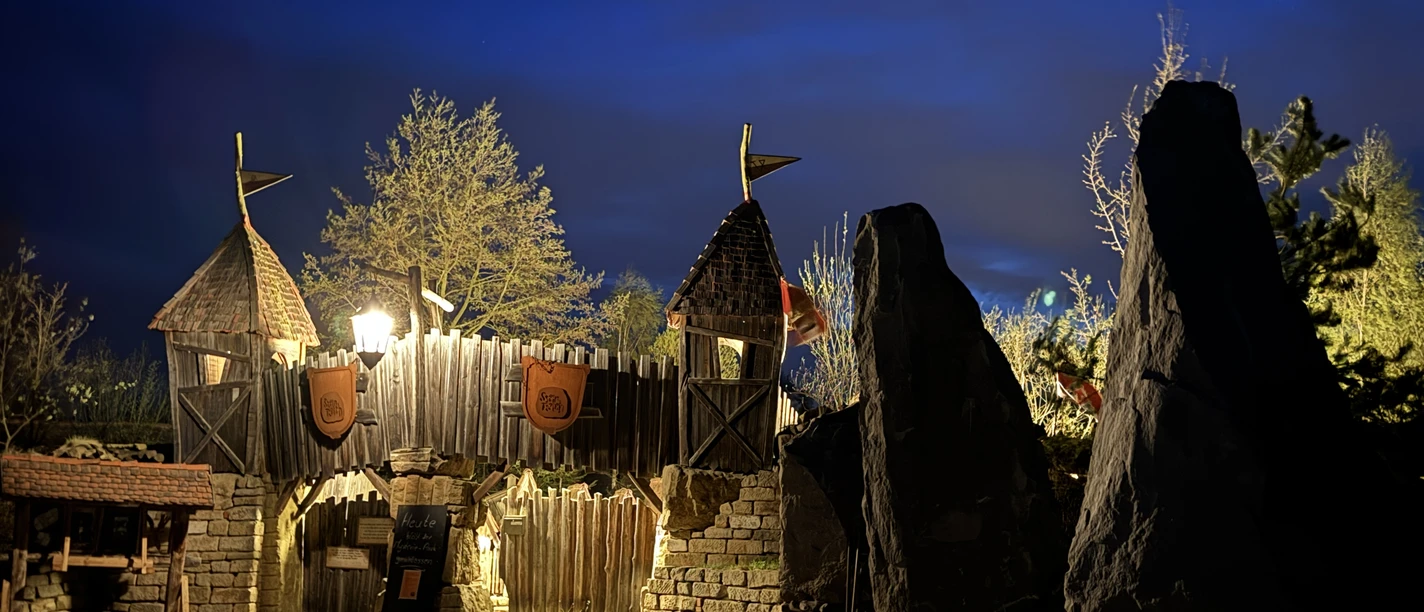 Bastei Erlebnis Steinreich Holztor mit Türmen und Fahnen, beleuchtet bei Nacht, umgeben von Bäumen und großen Steinen, vor einem tiefblauen Himmel.