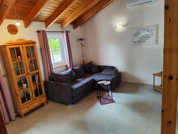 Wohnbereich des Ferienhauses Oelker Wohnzimmer mit grauem Ecksofa, linksseitig ein Buffetschrank aus Holz, Stehlampe, Fenster mit Vorhangschals