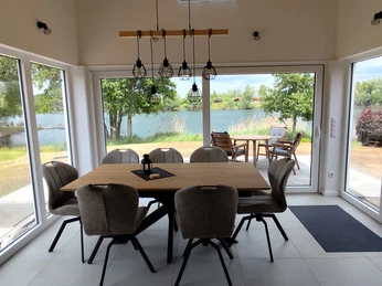 Essbereich des Ferienhauses Oelker Esstisch aus Holz mit sieben Polsterstühlen in Taupe, Hängelampe, komplette Fensterfront im Sitzbereich, Blick auf See