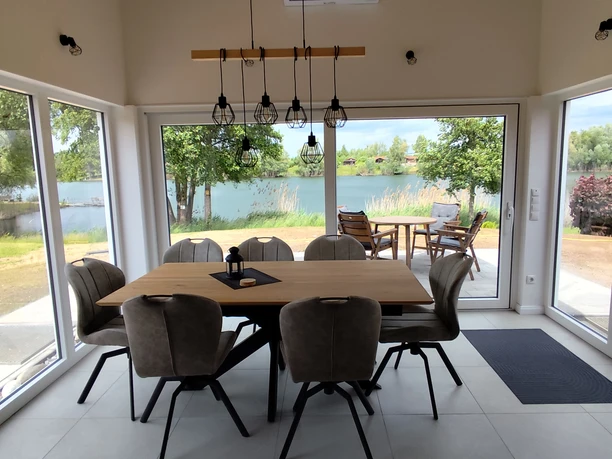 Essbereich des Ferienhauses Oelker Esstisch aus Holz mit sieben Polsterstühlen in Taupe, Hängelampe, komplette Fensterfront im Sitzbereich, Blick auf See