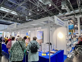 REHACARE Düsseldorf2024_Messestand2.jpg Messestand mit Besuchern auf der REHACARE Düsseldorf 2024. Broschüren und Hilfsmittel werden präsentiert.