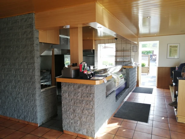 Modern gestalteter Imbiss mit offener Küche und Theke. Helle Fliesen und Holzdecke schaffen freundliche Atmosphäre.