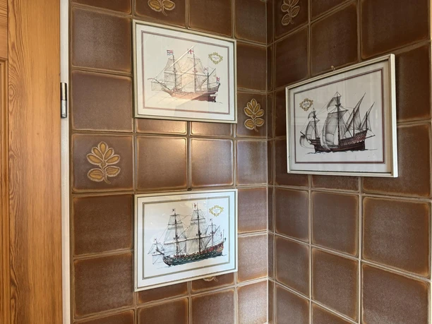 ferienwohnung-alscher-amelinghausen-retro-badezimmer-mit-Bilder.JPG Im Badezimmer der Ferienwohnung hängen drei gerahmte Bilder von historischen Segelschiffen an den Wänden mit braunen Fliesen. Die Fliesen haben ein Blumenmuster.