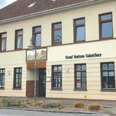Restaurant Brassierie Graf-Anton-Günther Das Bild zeigt das zweistöckige Gebäude "Graf Anton Günther" in einer urbanen Umgebung. Die Fassade ist in Beige gehalten und das Dach mit roten Ziegeln gedeckt. Um das Gebäude sind Straßen mit Verkehrsschildern und einem Fußgängerüberweg zu sehen.