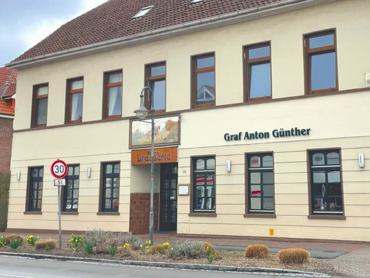 Restaurant Brassierie Graf-Anton-Günther Das Bild zeigt das zweistöckige Gebäude "Graf Anton Günther" in einer urbanen Umgebung. Die Fassade ist in Beige gehalten und das Dach mit roten Ziegeln gedeckt. Um das Gebäude sind Straßen mit Verkehrsschildern und einem Fußgängerüberweg zu sehen.