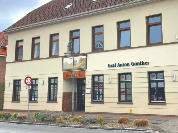 Restaurant Brassierie Graf-Anton-Günther Das Bild zeigt das zweistöckige Gebäude "Graf Anton Günther" in einer urbanen Umgebung. Die Fassade ist in Beige gehalten und das Dach mit roten Ziegeln gedeckt. Um das Gebäude sind Straßen mit Verkehrsschildern und einem Fußgängerüberweg zu sehen.