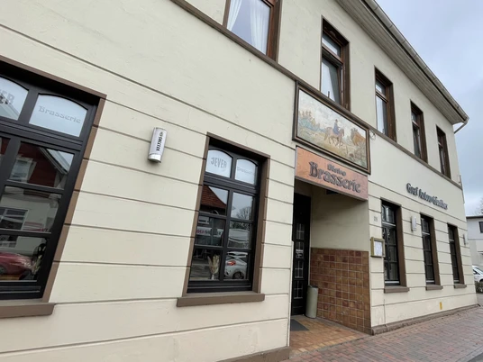 Restaunrant Brasserie Graf-Anton-Günther Das Bild zeigt die Außenfassade eines zweistöckigen Gebäudes, das als "Brasserie" ausgewiesen ist. Die Wände sind beige mit braunen Fensterrahmen. Über dem Eingang hängt ein farbiges Schild mit einem Pferdemotiv. Neben der Tür befindet sich ein Leuchtkasten mit der Aufschrift "Essen".