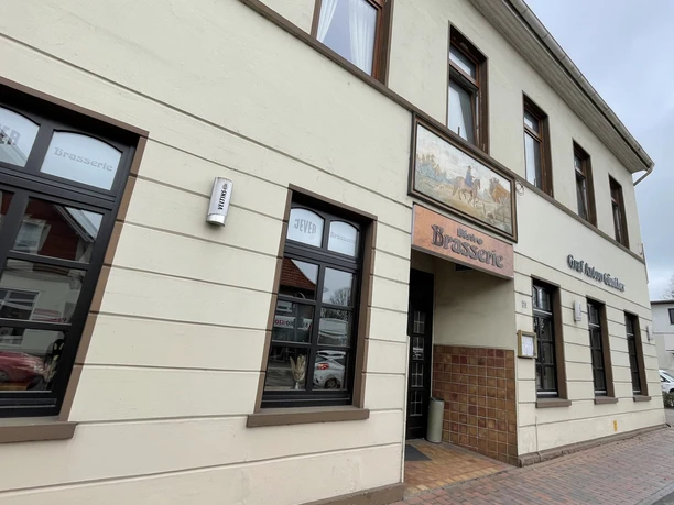 Restaunrant Brasserie Graf-Anton-Günther Das Bild zeigt die Außenfassade eines zweistöckigen Gebäudes, das als "Brasserie" ausgewiesen ist. Die Wände sind beige mit braunen Fensterrahmen. Über dem Eingang hängt ein farbiges Schild mit einem Pferdemotiv. Neben der Tür befindet sich ein Leuchtkasten mit der Aufschrift "Essen".
